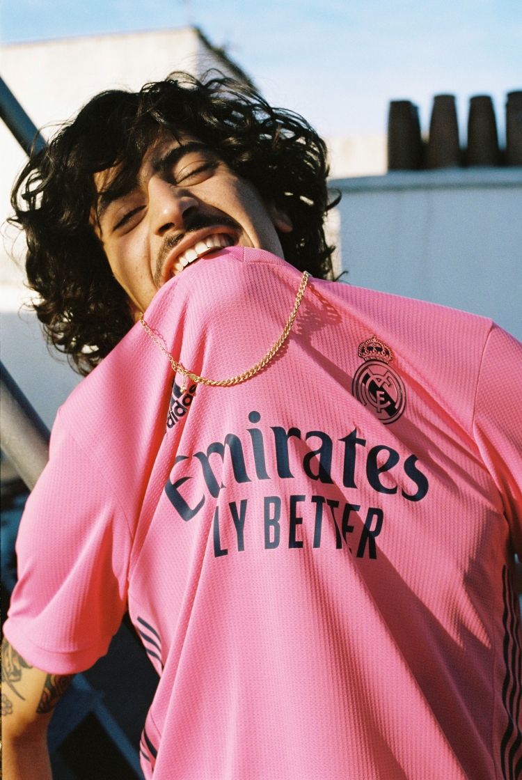 Young man in pink soccer jersey smiles on rooftop with bright sunlight. Gold chain necklace. Urban rooftop and sky background.