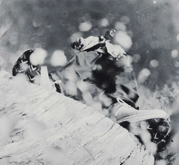 Black and white action shot of a person falling while wearing outerwear and sneakers on a rocky surface with bright bokeh.