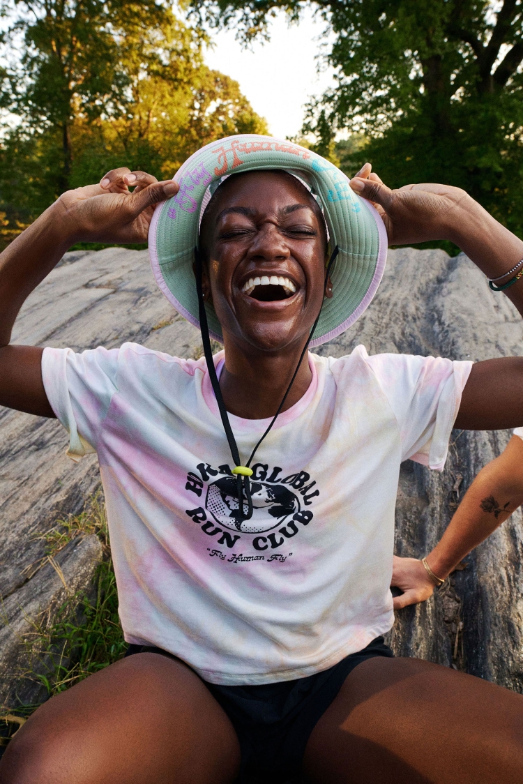 Smiling African-American woman in a bucket hat and t-shirt sits outdoors on a rock with hands on her head, trees visible in the background.