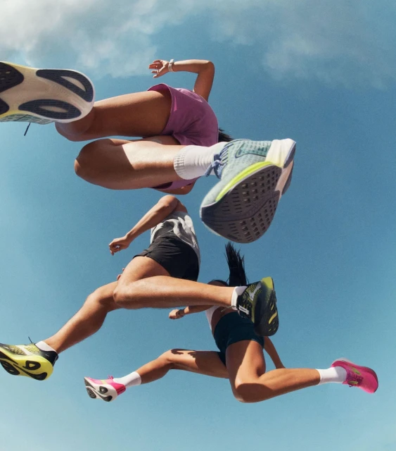 Three athletic women jump with running shoes against a blue sky in a low-angle shot that highlights their dynamic mid-air movement.