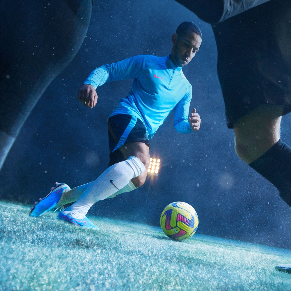 Male soccer player in blue uniform dribbles a multi-colored soccer ball on a grass field, illuminated by stadium lighting at night.