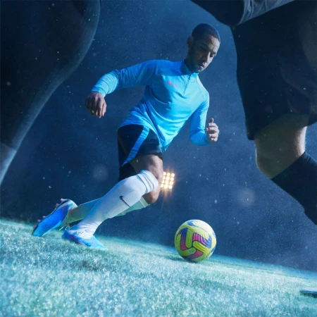 Male soccer player in blue uniform dribbles a multi-colored soccer ball on a grass field, illuminated by stadium lighting at night.