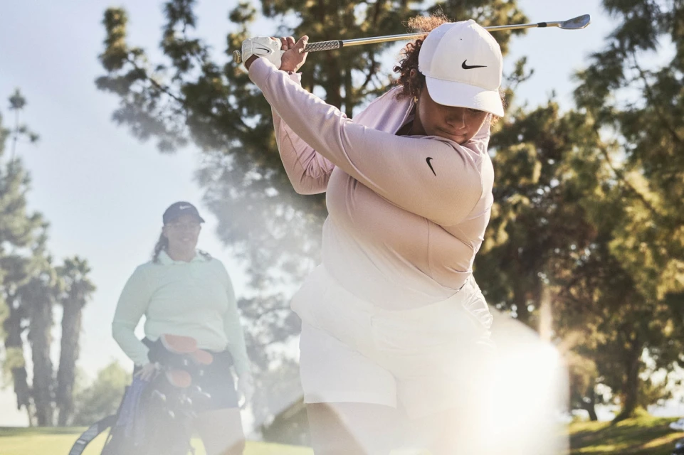 Woman in sportswear swinging a golf club on a sunny course with a companion; trees and blue skies provide a natural backdrop.