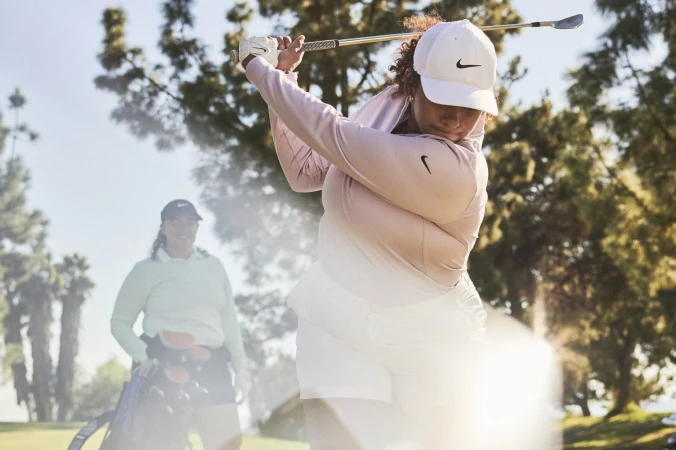 Woman in sportswear swinging a golf club on a sunny course with a companion; trees and blue skies provide a natural backdrop.
