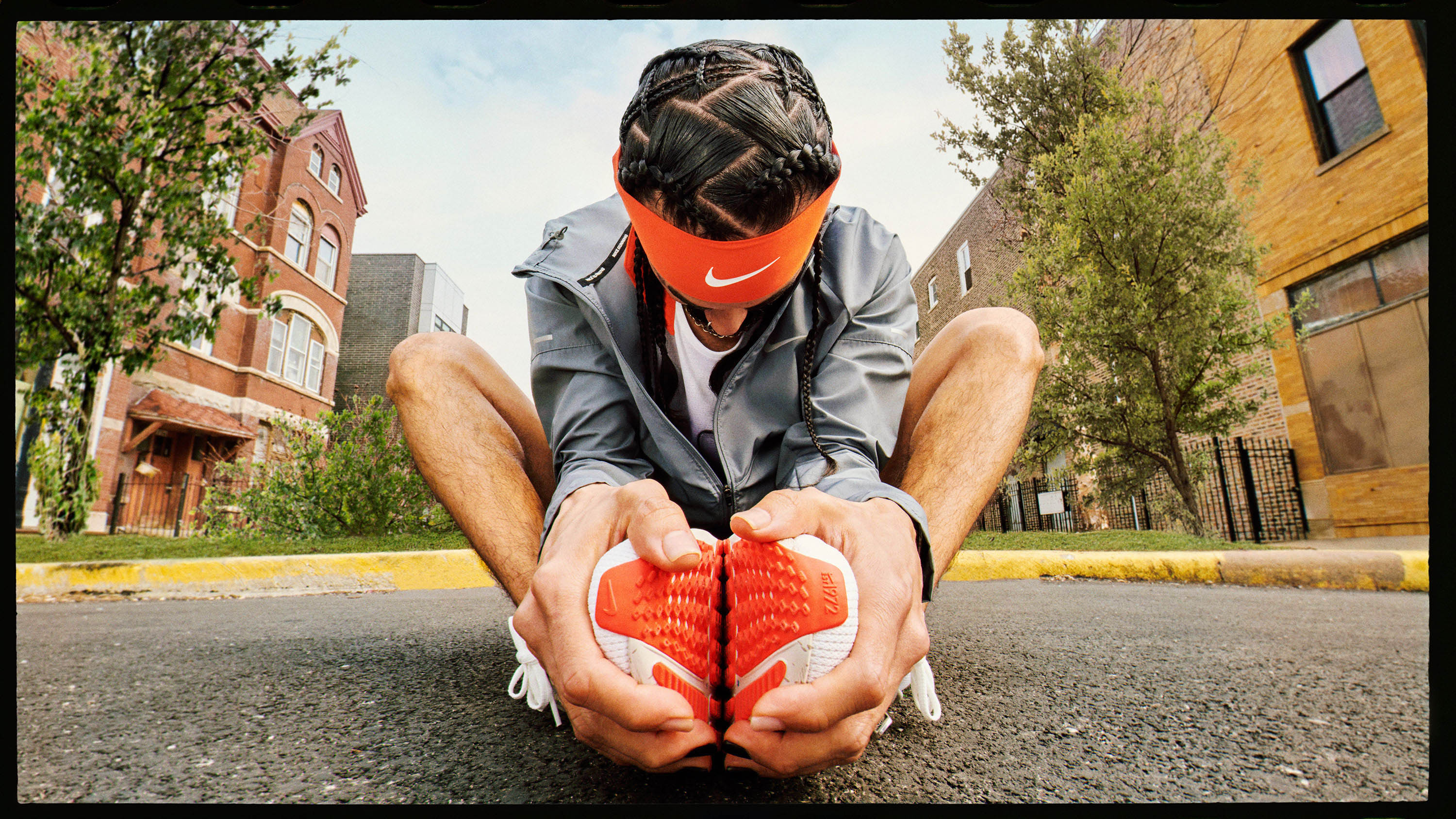 Low angle view of a man with an orange Nike headband sitting on pavement while holding his Nike running shoes against a city street backdrop.