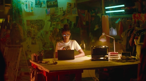Young adult female works at desk in cluttered room illuminated by a desk lamp and overhead lighting, focusing on her laptop.