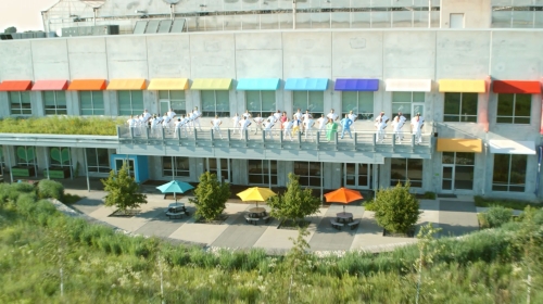 Aerial view of a modern building with colorful awnings where a group of adults in white outfits perform a synchronized activity.