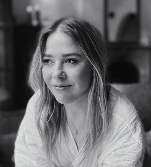 Black and white portrait of a woman with long blonde hair wearing a white textured shirt, gazing off to the side indoors.