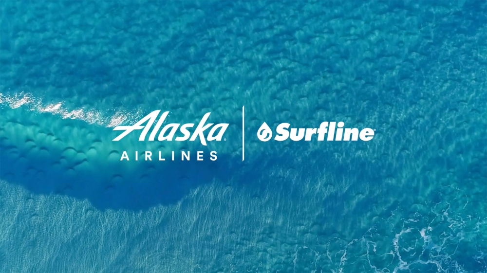 Aerial view of ocean water with wave and logos for Alaska Airlines and Surfline, emphasizing travel and partnership themes.