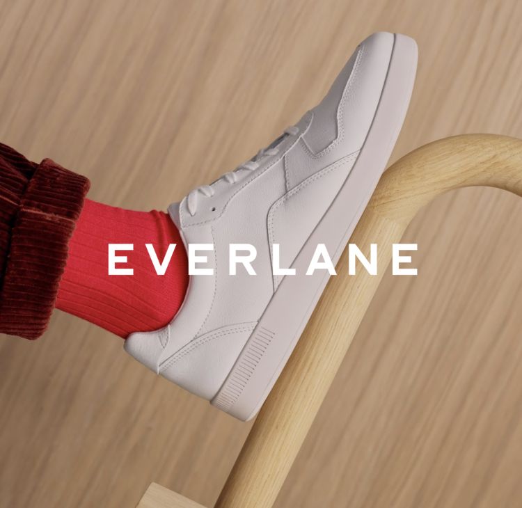 White sneaker on a wooden stool with a visible red sock and the EVERLANE logo. Close-up shot, product-focused, clean design.