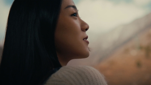Profile shot of an Asian woman with dark hair wearing a sweater, set against a blurred background featuring mountains.