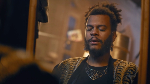 Portrait of a person with a beard and glitter makeup wearing a patterned jacket and necklace, looking towards the left.