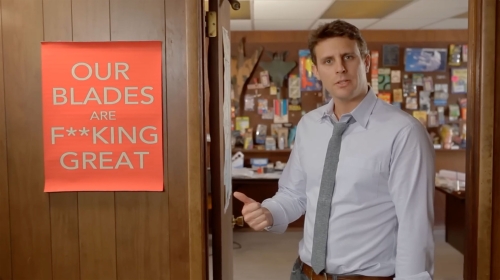 Adult male gestures toward orange poster reading 'Our Blades Are F***ing Great' in an indoor setting. 