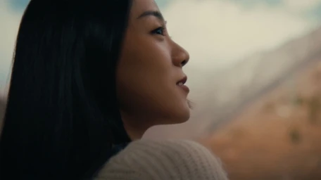 Profile view of an East Asian woman with long dark hair looking upward against a mountain landscape.