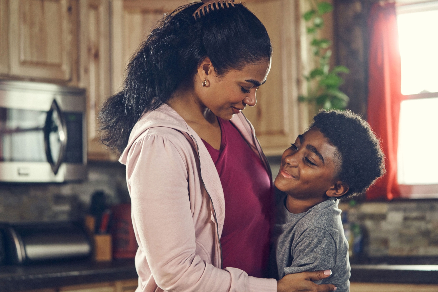 A mother and her son are standing in a kitchen. The mother is hugging her son, and they are both smiling at each other indoors.