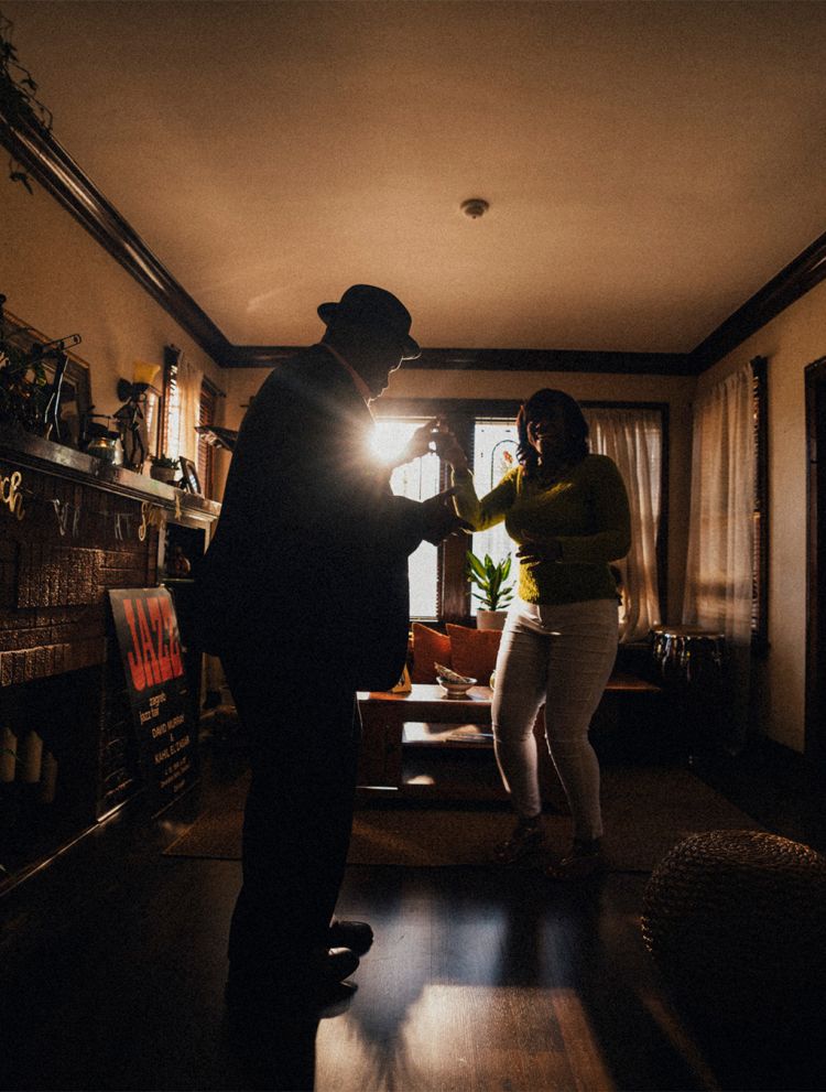 Couple dances in a sunlit living room. A man in a dark suit and hat leads a woman in white pants and a green sweater. Warm light fills the scene.
