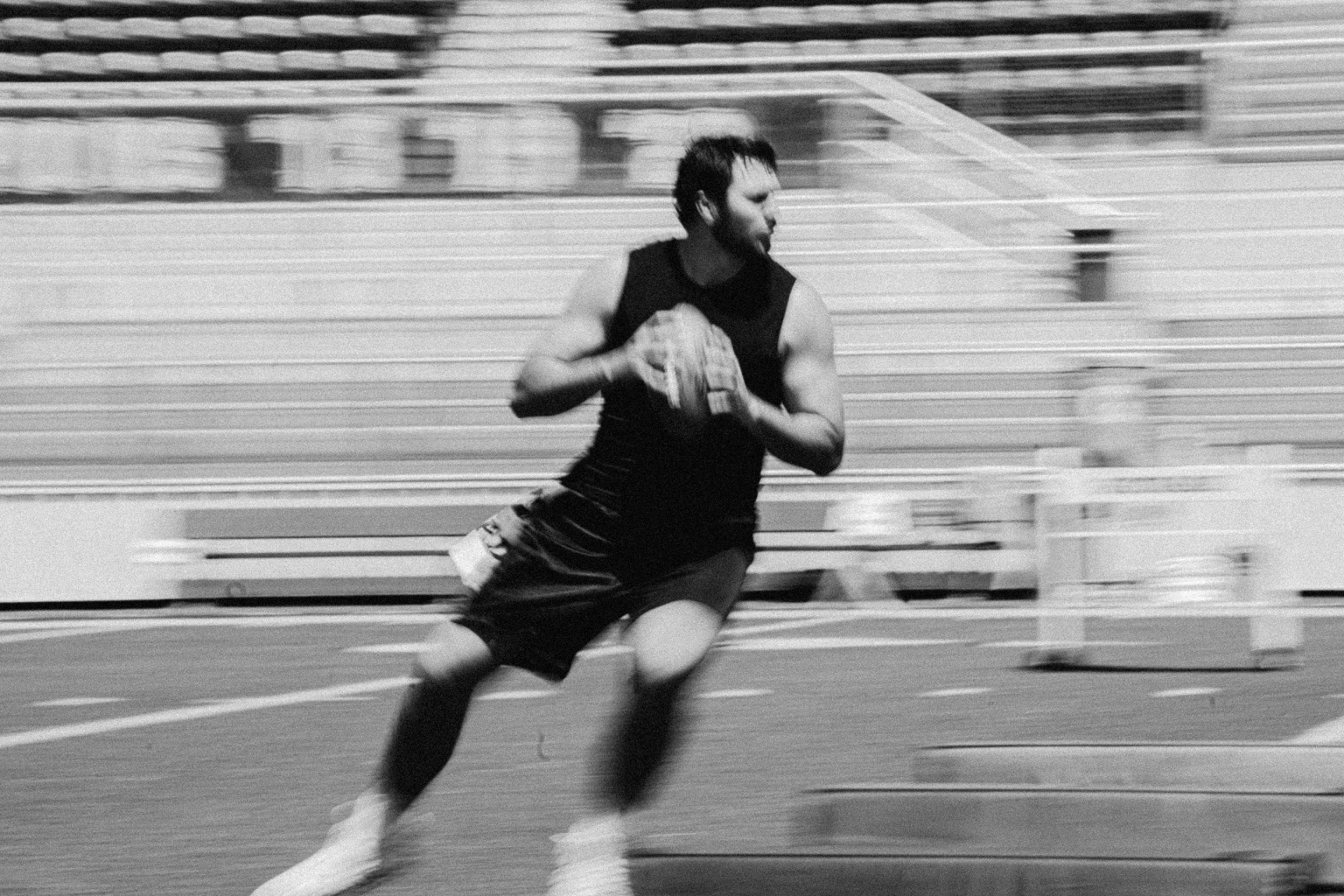 Monochrome action shot of an adult male running with a football on a field in front of stadium seating, motion blur effect.