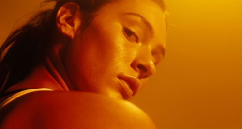 Close-up portrait of a woman with dark hair, gazing down, in warm, golden lighting that highlights the contours of her face and shoulder.