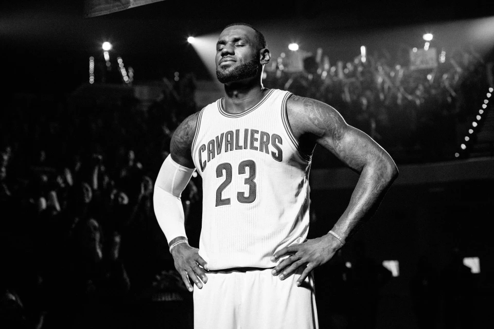 Monochrome shot of an adult male basketball player wearing a Cavaliers jersey, standing on a court in front of blurred crowd in a stadium.