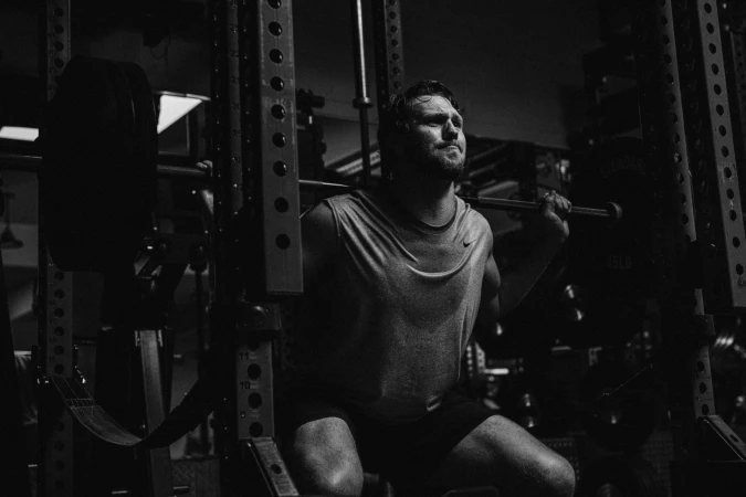 Adult male weightlifter doing squats with heavy barbell in black and white fitness photoshoot inside of a dark gymnasium.