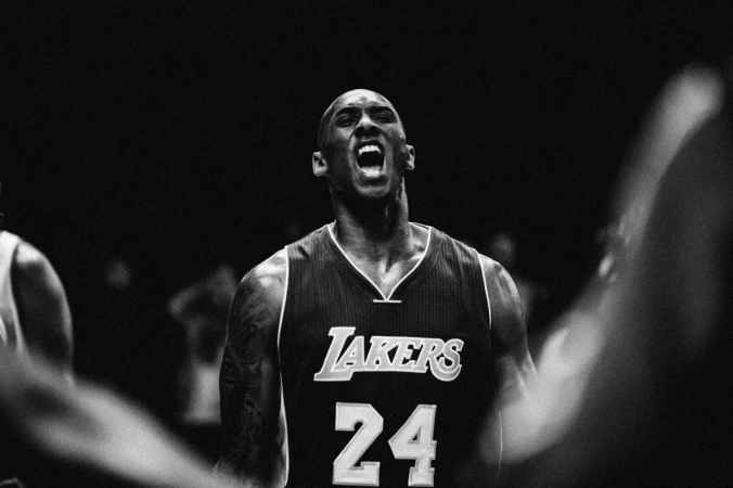 Close-up shot of an adult male basketball player in a Lakers jersey, face contorted while yelling; the scene is lit in black and white.