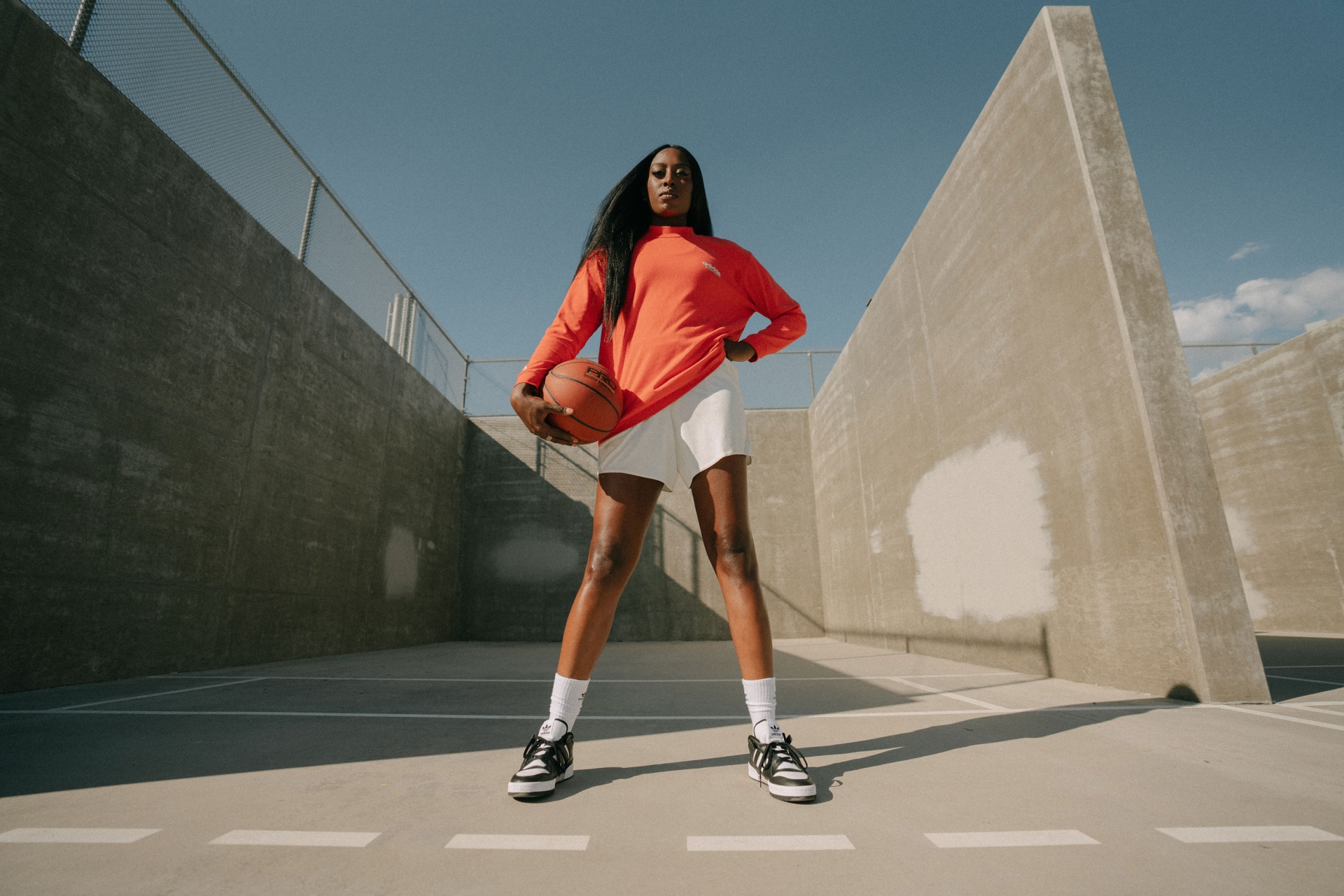 Athletic Black woman in sportswear poses on an outdoor court, holding a basketball against a concrete wall and sunny blue sky.