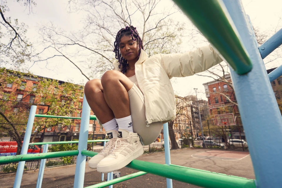Young woman in cream jacket, beige athletic wear, and sneakers sits on a green and blue jungle gym in a park setting outdoors.