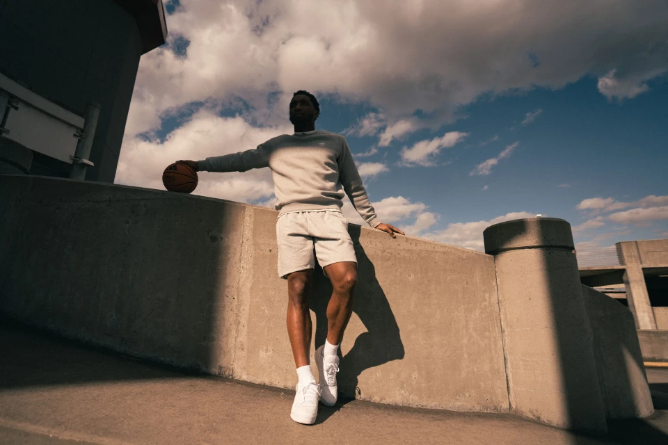 Athletic African-American man in sweatsuit stands on a concrete rooftop with blue sky, holding a basketball, wearing sneakers.