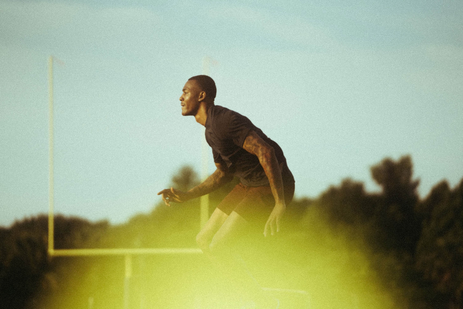 An adult male with tattoos does a leaping exercise outdoors in daytime with a focused expression. The background shows a football goal post.