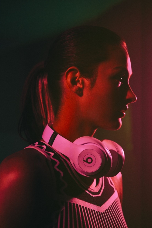 Profile shot of an adult female wearing a tank top and white Beats headphones, side lit with red and green gels in a dark studio.