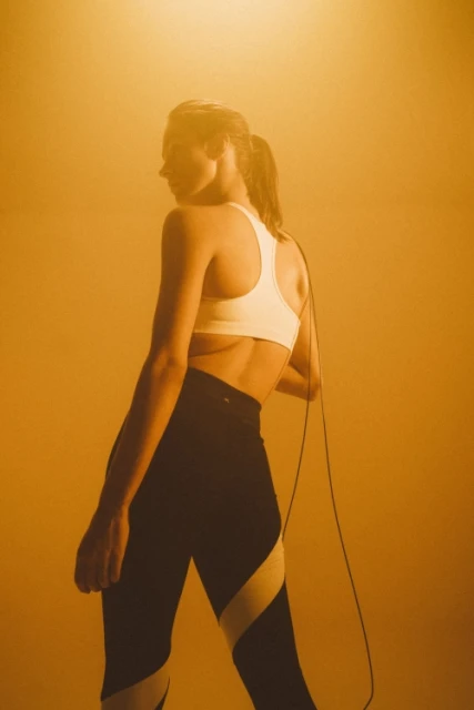 Athletic woman in sports bra and leggings, back view, stands in a studio with a jump rope around her neck under yellow lighting.