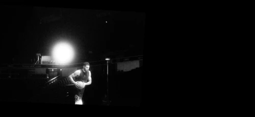 Black and white shot of a basketball player holding a ball on a dark indoor court, likely in a sports arena or gym.