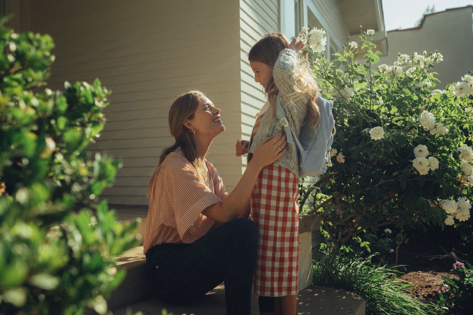 A mother and daughter share a tender moment in front of their home, with sunlight adding a warm glow to the scene.