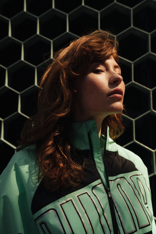 Portrait of a red-haired adult female in a teal and black Puma jacket against a dark honeycomb background, lit by natural side lighting.