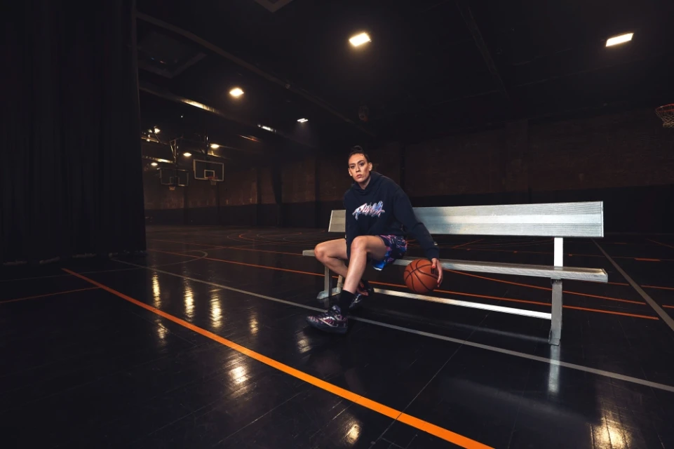 A young woman sits on a bench indoors in athletic wear, holding a basketball, with a basketball court visible in the background.