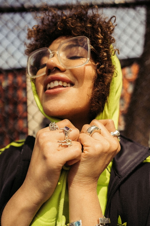 Stylish woman with curly hair smiles wearing glasses, rings, Adidas jacket, and neon green hoodie standing against a chain-link fence.