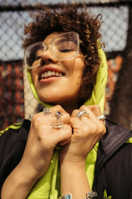 Stylish woman with curly hair smiles wearing glasses, rings, Adidas jacket, and neon green hoodie standing against a chain-link fence.