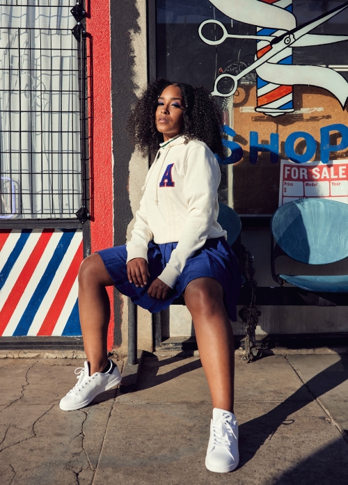 Woman with dark curly hair wearing a white hoodie and blue shorts sitting outside of a barber shop on a sunny day.