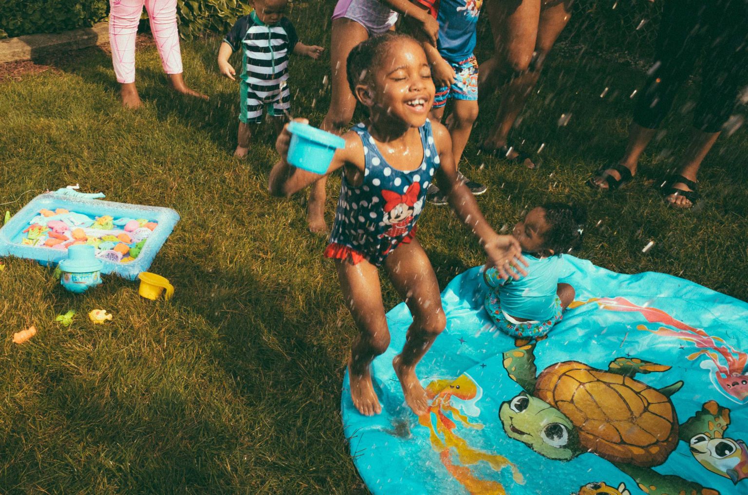 A group of children play outside on the grass with water, with a girl in a polka dot bathing suit smiling as she jumps.