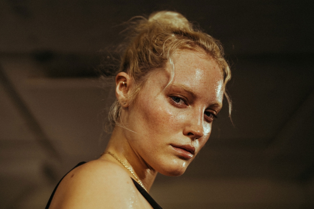 Close-up portrait of a sweaty blonde woman with a neutral expression wearing a tank top in a dimly lit indoor setting.