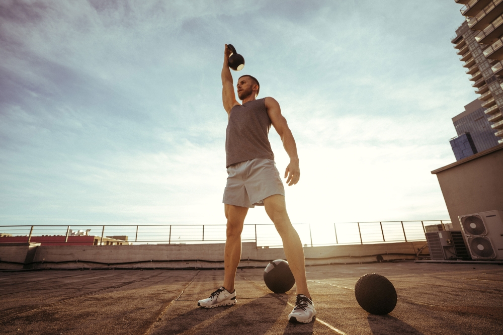 Athletic man lifting kettlebell on rooftop with medicine balls, set against a bright blue sky with city buildings in background.