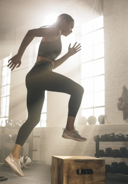 Athletic woman in fitness apparel powerfully jumps onto a wooden box in a brightly lit gym, showcasing strength and determination.