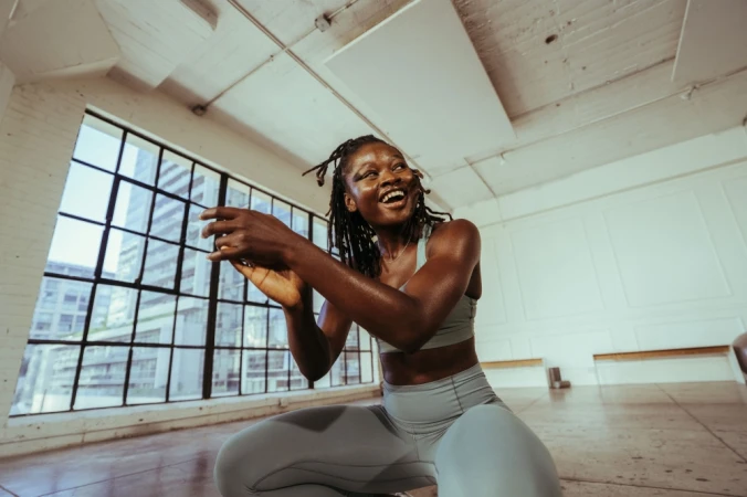Fitness portrait of a Black woman in a light blue athletic set crouching in a bright studio with large grid windows.