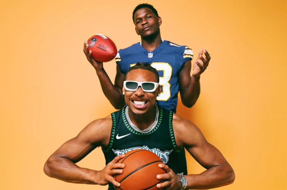 Two African American men pose with sports equipment in a studio: one holding a football and the other holding a basketball.