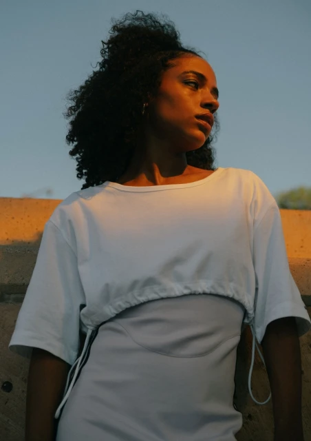 Portrait of an adult female with curly hair wearing a white top and a light-lavender dress against a clear sky with golden-hour light.