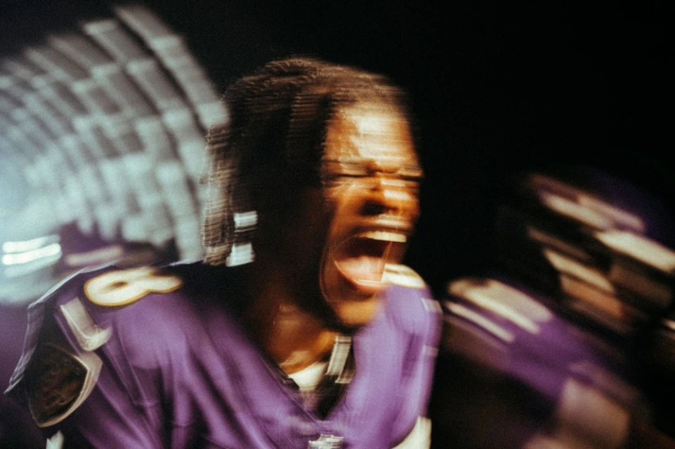 An African American athlete in a purple jersey shouts with visible excitement at night with blurred stadium lights in the background.