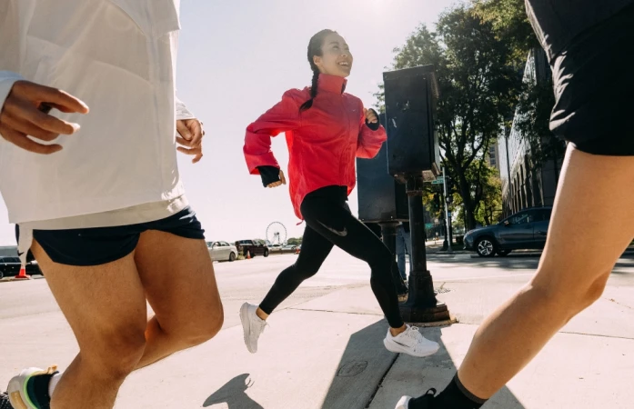 Three runners on a city street, focusing on a smiling East Asian woman in a bright pink jacket and black leggings.