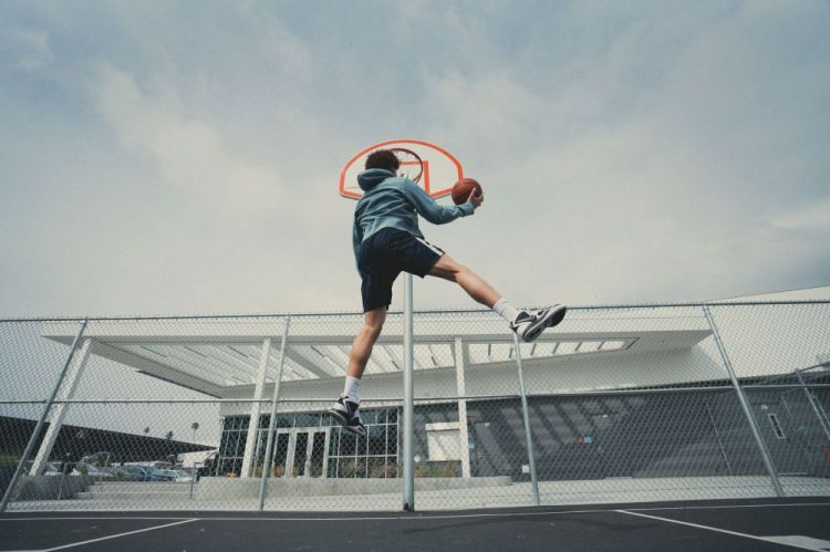 An athletic man in mid-air playing basketball, captured in a dynamic full shot on an outdoor court with a modern building backdrop.