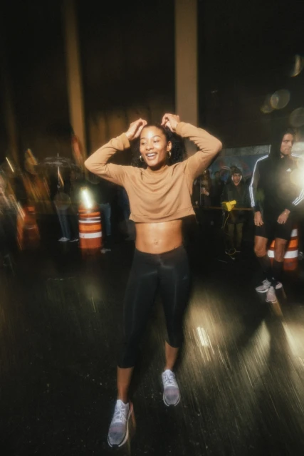 Woman smiles, placing hands on head in an urban street setting at night with blurred figures and orange traffic cones in background.