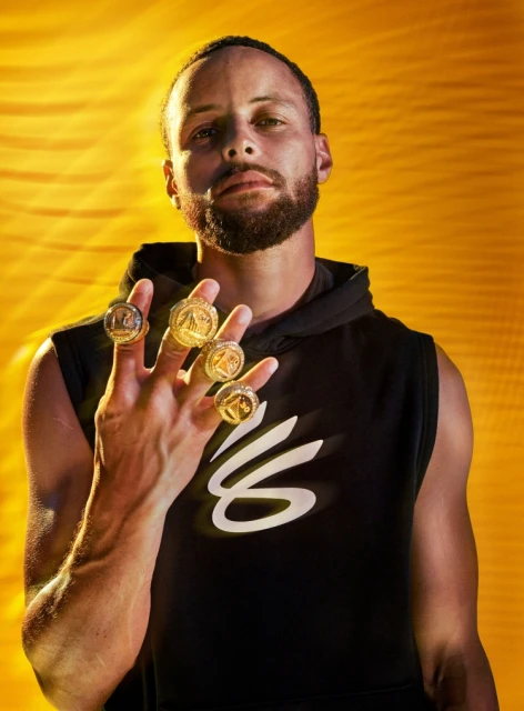 Adult male with beard wearing a black sleeveless hoodie showing off his championship rings against a warm yellow backdrop.